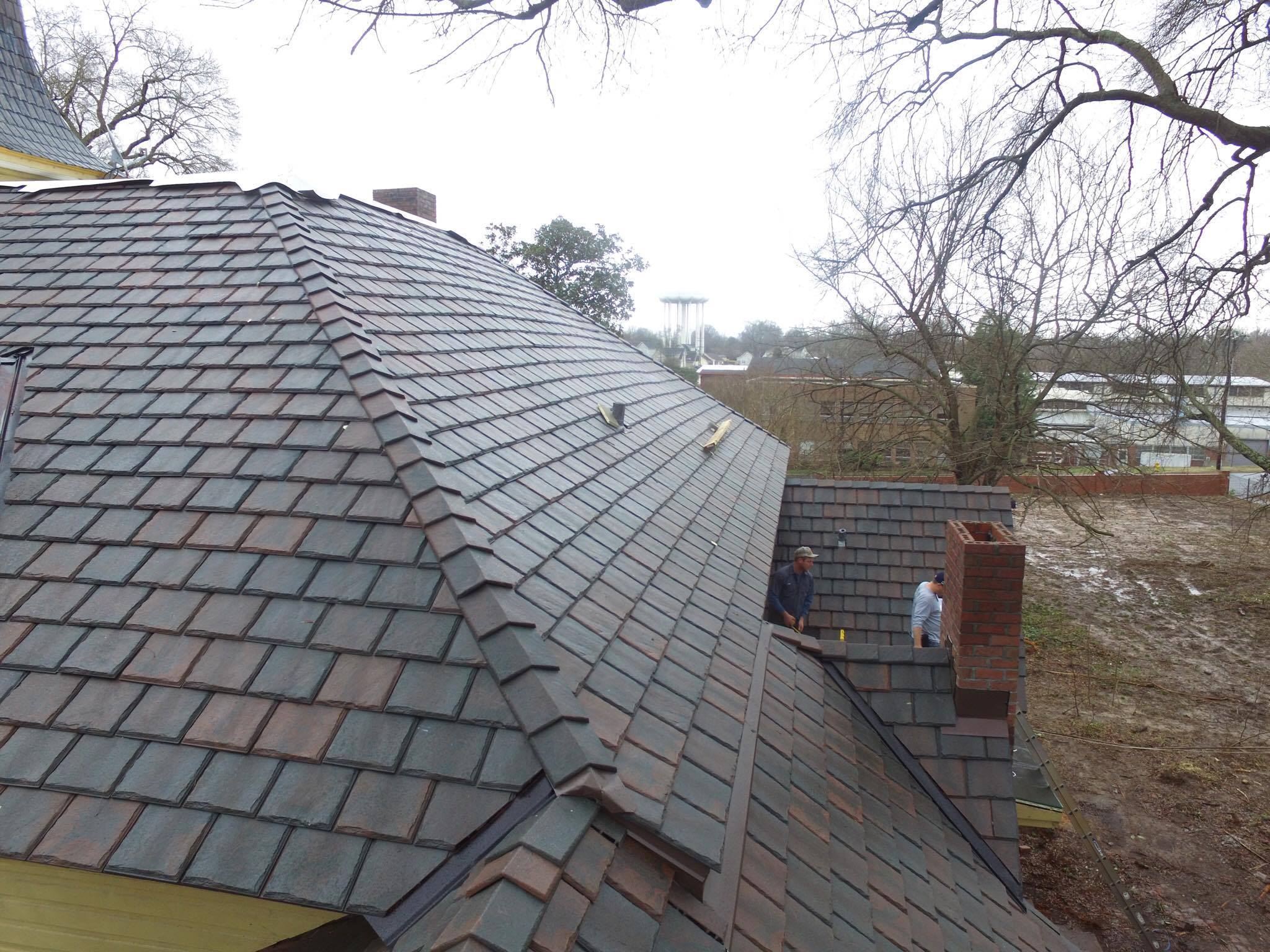 Aerial view of a Brava slate installation in progress in Statesville, NC, showing the dimensional slate profile, a brick chimney, and two installers working on a dormer.
