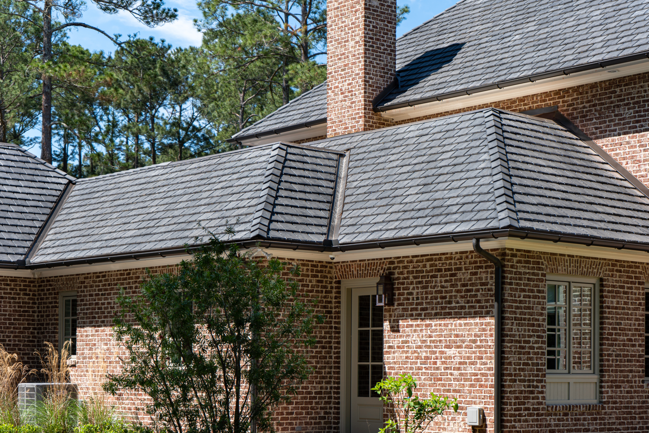 A traditional red brick estate home with a Brava Light Arendale slate roof, dark gutters, and tall longleaf pines in the background.