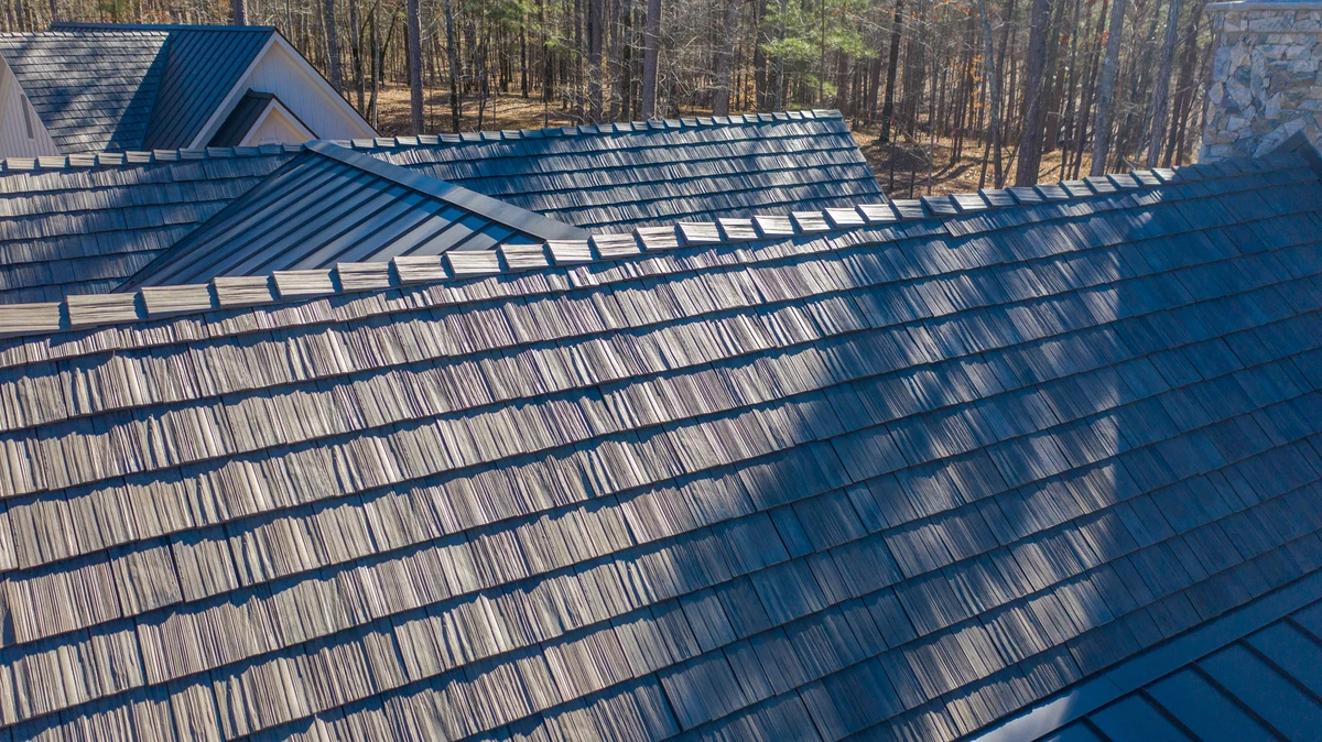 Aerial view of a Brava Cedar Shake roof in a weathered gray finish set against tall pine and hardwood trees with a stone chimney in the background.