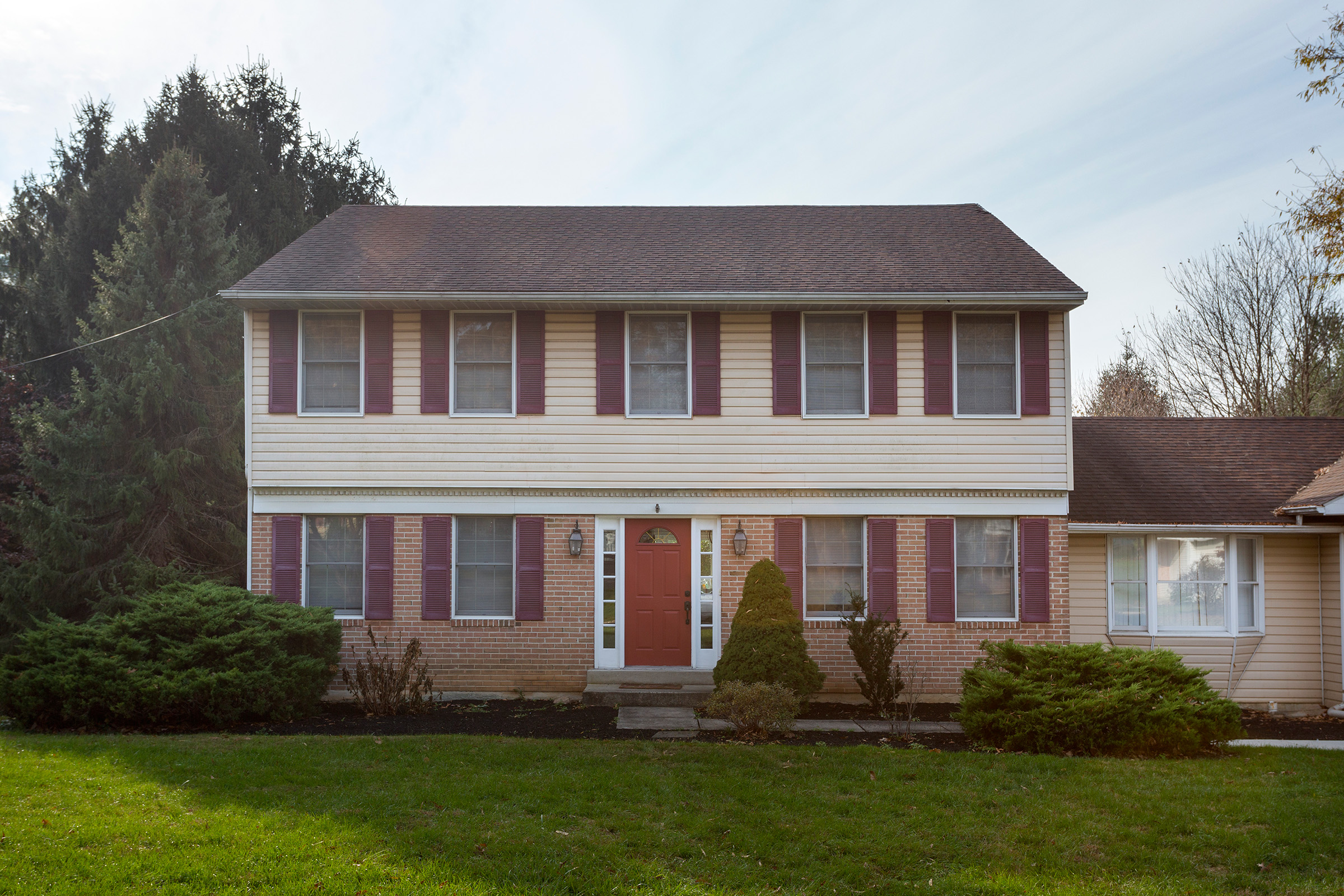 A 1990s two-story colonial home with faded beige vinyl siding, burgundy shutters, brick first floor, and a red front door.